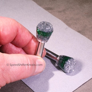 Pode incluir: Dois manípulos de mudança de velocidades com um eixo metálico cilíndrico e uma parte superior arredondada. A parte superior é de material transparente com purpurinas prateadas e uma banda horizontal de purpurinas verdes. Texto: SpeedShifterKnobs.com.