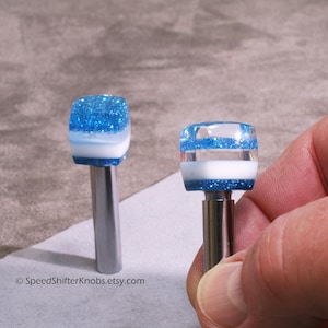 May include: Two custom shift knobs with a blue glitter and white design. The knobs have a rectangular shape and are mounted on a silver metal shaft. The Etsy shop name "SpeedShifterKnobs.etsy.com" is visible in the image.