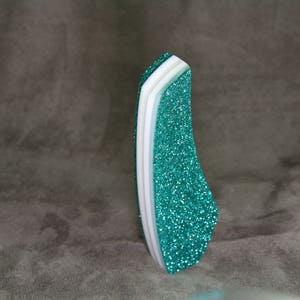 Peut inclure: Une lime à ongles pailletée turquoise avec des couches blanches et turquoise clair. La lime à ongles est incurvée et a une finition scintillante. Le fond est gris uni.