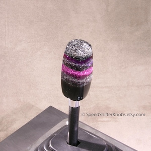May include: A custom shift knob with a black base and a glittery top. The top section features horizontal bands of silver, purple, and black glitter. The shift knob is mounted on a black metal rod.