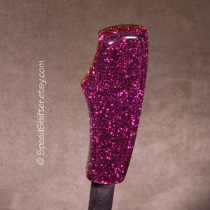 Op de afbeelding: Een aangepaste auto versnellingspookknop met een levendige magenta glitter afwerking. De knop heeft een gebogen, ergonomisch ontwerp en een glanzend, gepolijst oppervlak. De bovenrand heeft een roodgouden rand. De tekst "SpeedShifter.etsy.com" is zichtbaar.