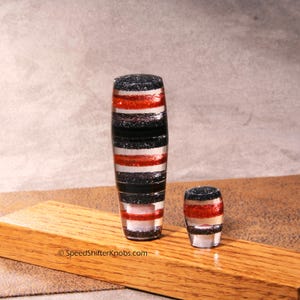 May include: Two cylindrical gear shift knobs featuring horizontal bands of black and red glitter embedded in a clear resin material. The taller knob is on the left and the shorter knob is on the right. The text SpeedShifterKnobs.com is visible.