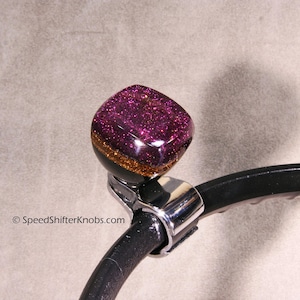 以下が含まれることがあります： キラキラ光る紫色のトップと金と黒のアクセントが付いた、カスタムカーのシフトノブ。ノブは、クロム色のクランプが付いた黒い金属アームに取り付けられています。