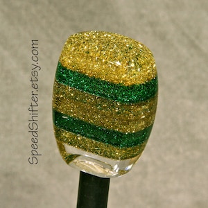 May include: A custom car shift knob with a clear base and a glittery design. The knob features alternating horizontal stripes of gold and green glitter. The text "SpeedShifter.etsy.com" is printed vertically on the left side.
