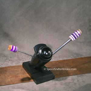 May include: A black, industrial-style object with two arms extending outwards. The arms have decorative ends with purple and white stripes. The object is mounted on a black base. The text "SpeedShifterKnobs.com" is visible.