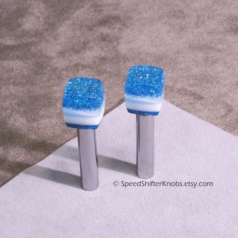 May include: Two custom shift knobs with a blue glitter top, white and blue layers, and a silver metal stem. The knobs are designed for a car's gear shift lever. The Etsy shop name SpeedShifterKnobs.etsy.com is visible.