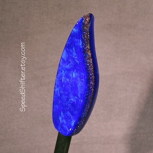 May include: A blue, leaf-shaped hair stick with a gold glitter accent along one side. The hair accessory has a glossy finish and is held by a dark, thin stick. The text "SpeedShifter.etsy.com" is printed on the left side.