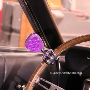 Op de afbeelding: Een aangepaste stuurknop met een paarse glittertop en chromen basis. De knop is gemonteerd op een houten stuurwiel. De afbeelding is gemaakt in een voertuig.