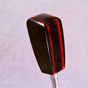 Puede incluir: Un pomo de palanca de cambios negro y rojo con forma rectangular y acabado pulido. El pomo tiene dos finas rayas rojas que recorren verticalmente los lados. El pomo está unido a una varilla metálica plateada.
