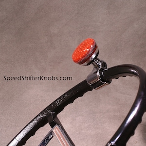 Op de afbeelding: Zwart stuurwiel met een rode glitter draaiknop. De draaiknop heeft een chromen basis. De tekst "SpeedShifterKnobs.com" is zichtbaar. Interieur van een voertuig.