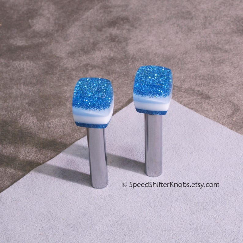 May include: Two shift knobs with a blue glitter top, a white stripe, and a silver metal stem. The knobs are displayed on a white surface. The text "&copy; SpeedShifterKnobs.etsy.com" is visible.