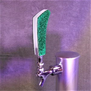 Puede incluir: Un tirador de cerveza con un diseño curvo. El tirador presenta una superficie verde brillante y brillante enmarcada por un borde blanco y una base de metal plateado. El grifo está unido a un cilindro de metal plateado.