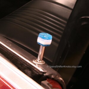 May include: A custom shift knob with a blue and white design. The knob is mounted on a silver metal rod and is set against a black car interior. The top of the knob has a glittery texture.