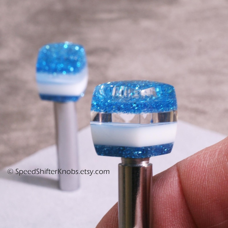 May include: Two custom shift knobs with a blue glitter top, white and light blue stripe, and a silver base. The knobs are designed for a car's gear shift lever. The Etsy shop name SpeedShifterKnobs.etsy.com is visible.