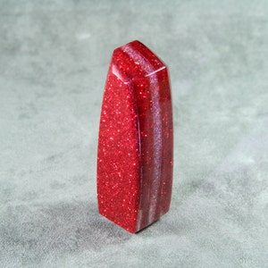 Può includere: Un oggetto decorativo a forma di obelisco con glitter rossi. L'oggetto ha una parte superiore sfaccettata e una base rettangolare. La superficie è ricoperta di glitter rossi, con strisce verticali di un colore rosso più chiaro. L'articolo è in piedi su una superficie grigia.