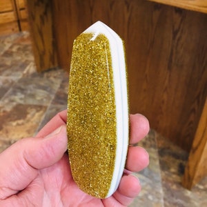 Puede incluir: Un obelisco dorado con purpurina y detalles blancos y dorados. El obelisco se sostiene en una mano y tiene forma rectangular con la parte superior puntiaguda. La purpurina dorada cubre la mayor parte del obelisco.