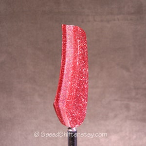Op de afbeelding: Een rode, met glitter bedekte versnellingspookknop. De versnellingspookknop heeft een levendige rode kleur met sprankelende glitter. De vorm is gebogen en langwerpig, met een gladde, gepolijste afwerking. De Etsy shopnaam, SpeedShifter.etsy.com, is onderaan zichtbaar.