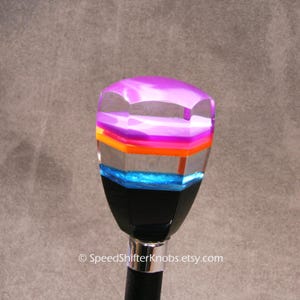 May include: A colorful shift knob with a clear, faceted top. The knob features layers of purple, orange, pink, and blue. The base is black and the shaft is silver and black. The Etsy shop name is visible at the bottom.