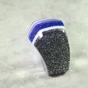 Peut inclure: Une bague épaisse avec un design en couches. Le dessus présente une bande violette, suivie d'une couche bleue scintillante, d'une bande blanche et d'une base noire pailletée. La bague a une forme incurvée et une finition brillante.