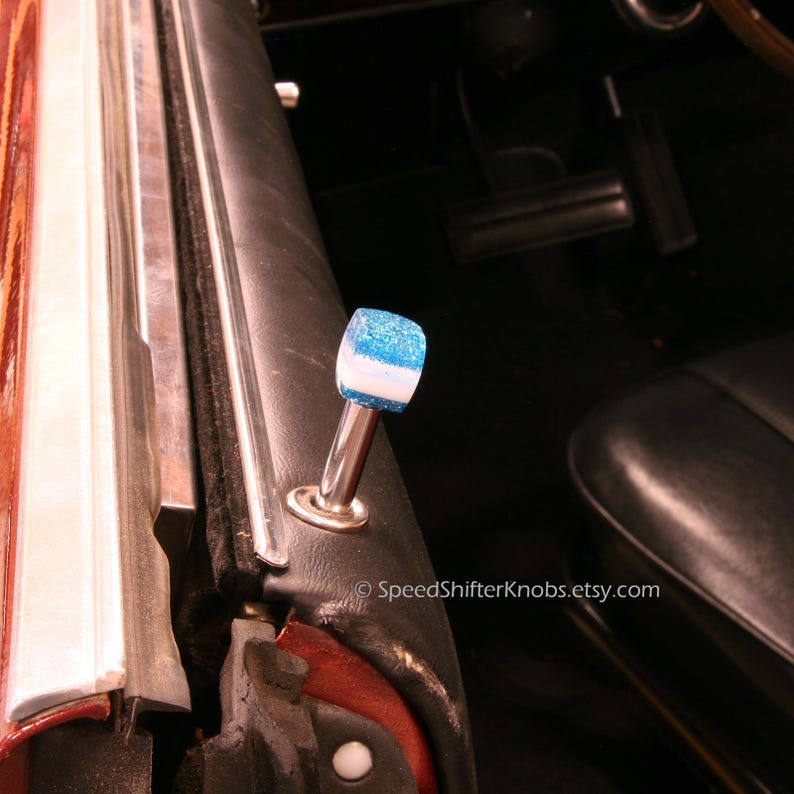 May include: A custom car interior detail featuring a blue and white glitter shift knob. The shift knob is mounted on a chrome stem and is set against a black interior.