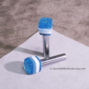 May include: Two blue and white glitter shift knobs with a silver metal stem. The knobs are square-shaped with a blue glitter top, a white stripe, and a silver stem. One knob is lying down, and the other is standing up. The Etsy shop name is visible.