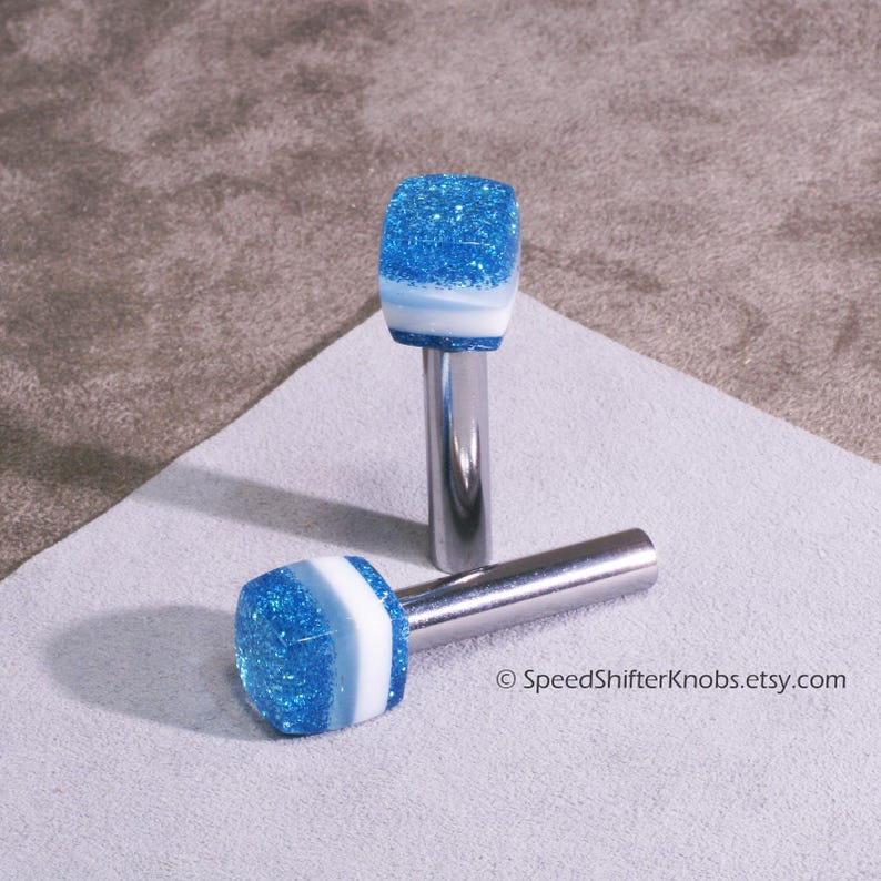 May include: Two blue and white glitter shift knobs with a silver metal stem. The knobs are square-shaped with a blue glitter top, a white stripe, and a silver stem. One knob is lying down, and the other is standing up. The Etsy shop name is visible.