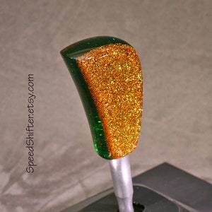 May include: A custom car shift knob with a green and gold glitter design. The shift knob has a curved shape and a metallic silver base. The text "SpeedShifter.etsy.com" is printed vertically on the side.