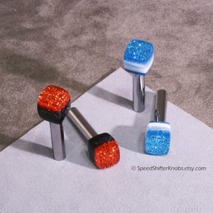 May include: Four glitter-topped shift knobs in red, black, and blue. The knobs have a rectangular shape and are mounted on silver metal stems. The image is on a gray surface. The text "SpeedShiftKnobs.etsy.com" is visible.