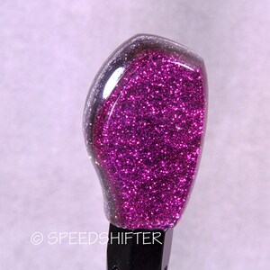 May include: A custom car shift knob with a vibrant purple glitter design encased in clear resin. The top portion is a deep, sparkling magenta, transitioning to a clear edge. The base is black, and the text "© SPEEDSHIFTER" is visible.