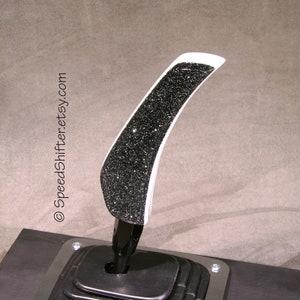 Op de afbeelding: Een aangepaste autoversnellingspook met een zwarte glitter bedekte grip en een witte rand. De pook is gemonteerd op een zwarte basis met een zwarte rubberen hoes. De tekst "SpeedShifter.etsy.com" is zichtbaar.