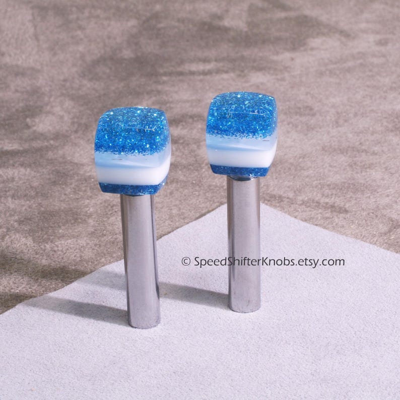 May include: Two custom shift knobs with a blue glitter top, white middle section, and a silver metal base. The knobs are designed for a car's gear shift lever. The blue glitter adds a decorative touch to the car's interior.