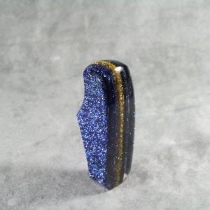 Könnte beinhalten: Ein blauer Sandstein-Edelstein mit Goldflocken und einem vertikalen Goldstreifen. Der polierte Stein hat eine einzigartige Form und ist vor einem grauen Hintergrund platziert. Der Edelstein ist ein dekorativer Gegenstand.