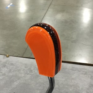 May include: A custom orange shift knob with a curved design. The knob features a vertical stripe of black glitter and a brown stripe. The shift knob is mounted on a chrome-coloured metal rod.