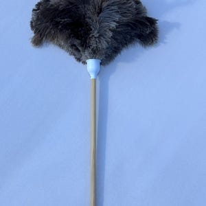 Könnte beinhalten: Ein grauer Staubwedel mit einem weißen Kunststoffgriff und einem hellbraunen Holzstab. Die Federn des Staubwedels sind dunkelgrau und flauschig, und der Griff ist mit einem weißen Kunststoffteil am Stab befestigt. Der Staubwedel ist zum Reinigen bestimmt.