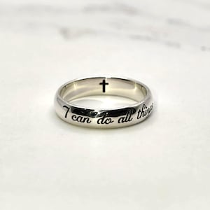 May include: A silver ring with the inscription "I can do all things" and a small cross. The ring is made of a shiny, reflective metal and is set against a white marble background. The ring is a piece of jewellery.