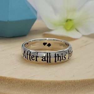 以下が含まれることがあります： 「After all this time」という刻印と2つの黒いハートがついた銀の指輪。指輪は木製の台の上に置かれています。