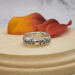 May include: A silver band ring with the inscription "Omnia Parata" in black lettering.