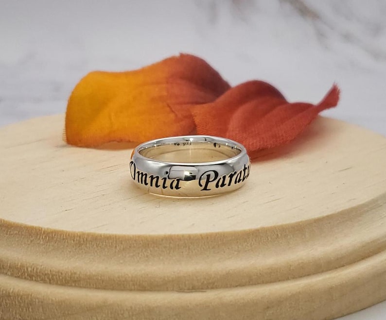 May include: A silver band ring with the inscription "Omnia Parata" in black lettering.