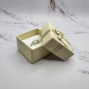 May include: A small, cream-colored ring box with a satin ribbon and bow. The box is open and a silver ring is inside.