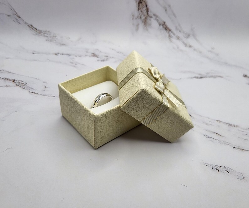 May include: A small, cream-colored ring box with a satin ribbon and bow. The box is open and a silver ring is inside.