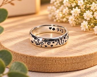 May include: A silver ring featuring the engraved text "We're All" along the outer band, accented with small card suit symbols. The interior of the band is stamped with "925" and additional card suit icons, including a heart and a spade.
