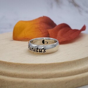 May include: A silver ring with the words "Paratus's" engraved on it. The ring is on a wooden surface with a fall-colored flower petal behind it.