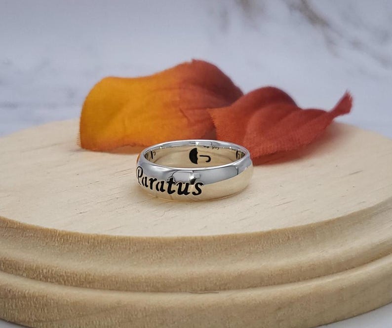 May include: A silver ring with the words "Paratus's" engraved on it. The ring is on a wooden surface with a fall-colored flower petal behind it.