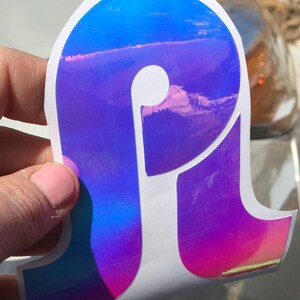 May include: A holographic sticker with a letter P in a bold, rounded font. The sticker is iridescent and shifts colors from blue to purple to pink.