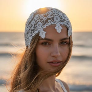 Può includere: Una cuffia in pizzo bianco caratterizzata da intricati ricami floreali e bordi smerlati che seguono il profilo della fronte. Il copricapo è realizzato in un delicato tessuto a rete trasparente con motivi dettagliati su tutta la superficie.