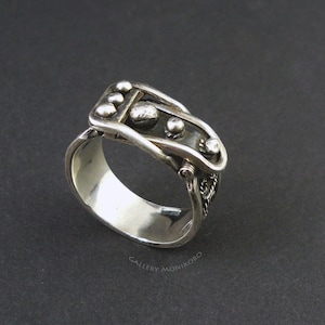 Può includere: Un anello d'argento con un designo di fibbia unico. La fibbia è dettagliata con piccole sfere d'argento e una superficie testurizzata. L'anello è realizzato in argento sterling.