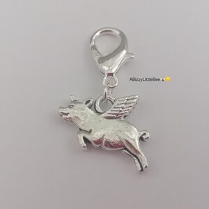 May include: A silver charm depicting a pig with wings, suspended from a clasp. The pig is shown mid-flight, with detailed wings and a textured body. The charm is suitable for a bracelet or necklace.