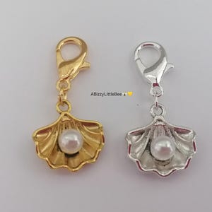 May include: Two gold and silver charms in the shape of a seashell with a pearl inside. The charms have a lobster clasp.