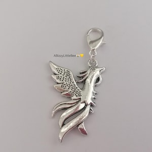May include: Silver charm featuring a detailed phoenix design with spread wings. The charm has a lobster clasp for attaching to a bag, keychain, or other accessories.