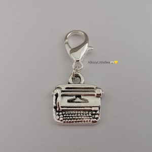May include: Silver typewriter charm with a lobster clasp. The charm has a detailed miniature typewriter design, including keys and a carriage. The text "ABizzyLittleBee" is visible in the upper right corner.